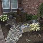 3 pillar stone water feature in front of house window with rocks and plants